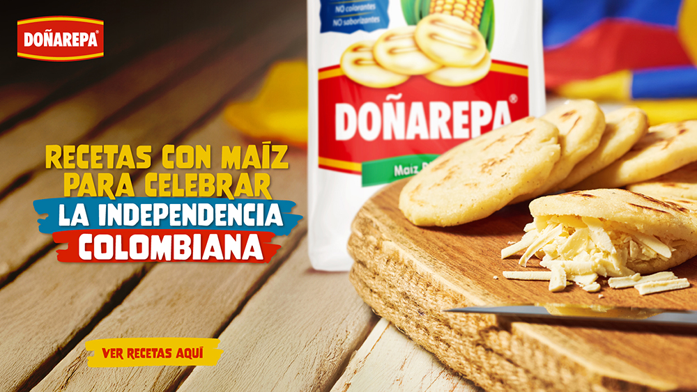 Vista de varias arepas de maíz rellenas con queso rallado sobre una tabla de madera. Al fondo, una tela de colores y contenedores con especias.